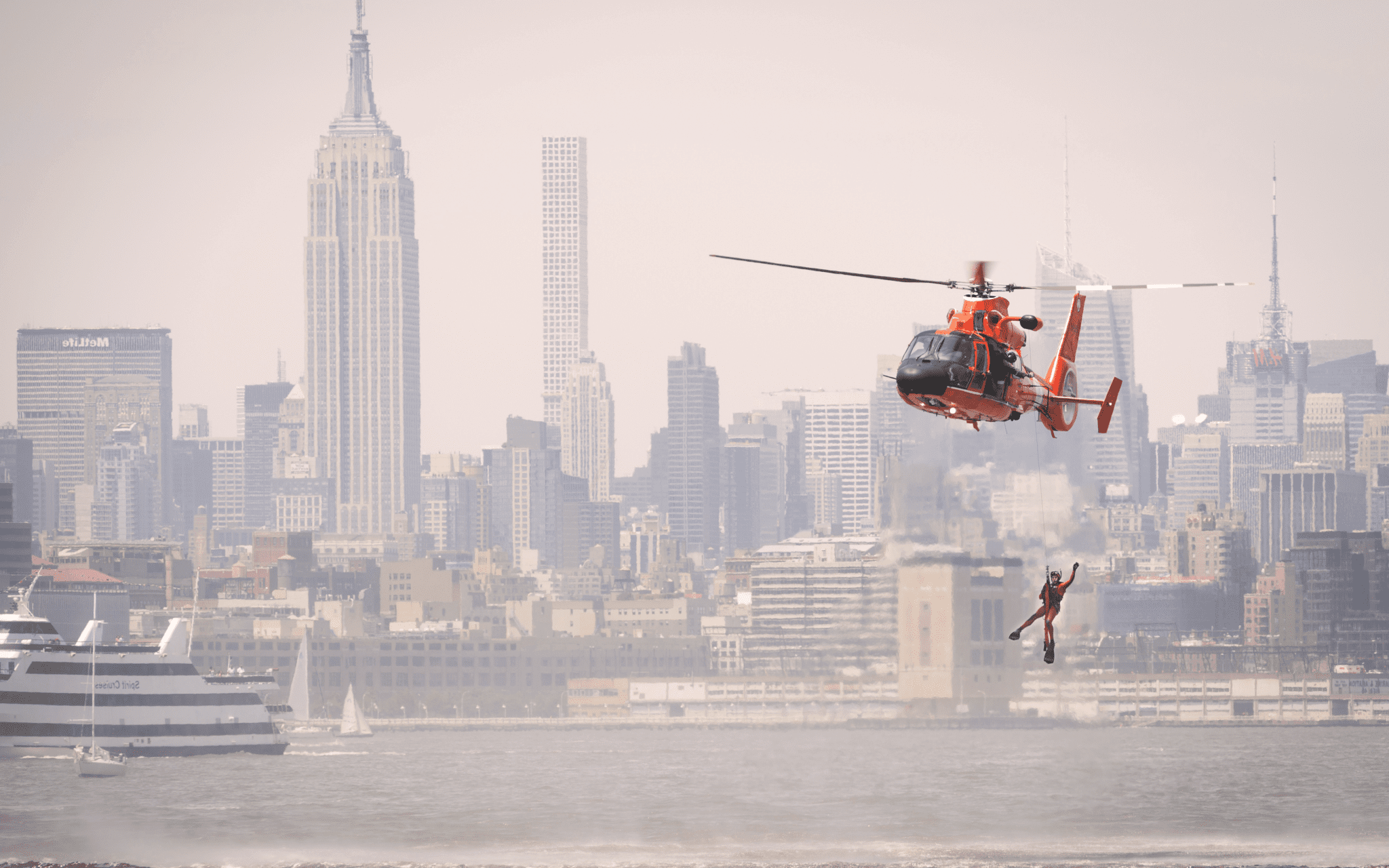 Coast guard member hangs from helicopter above the ocean in front of a city.