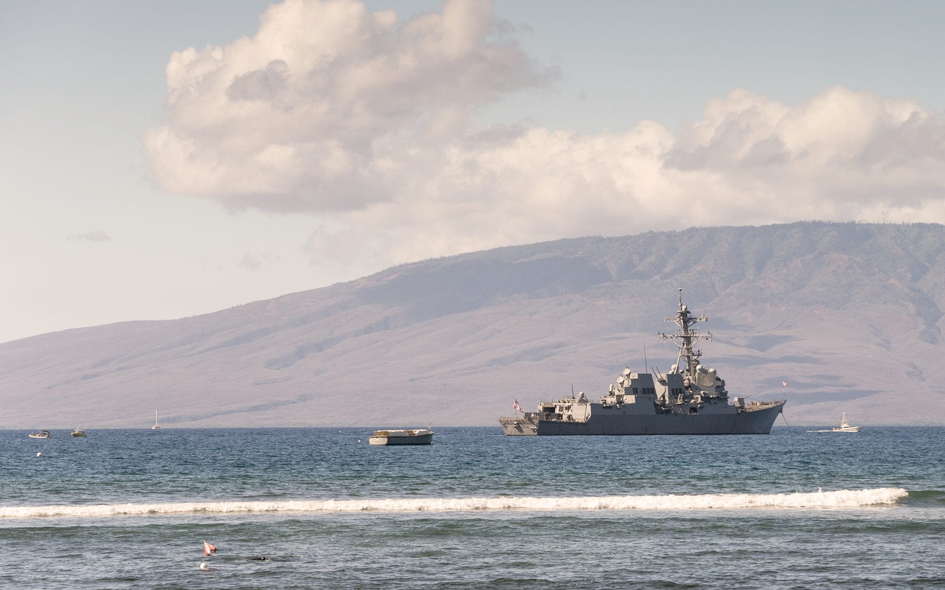 A Navy ship sits in the ocean in front of a mountain.