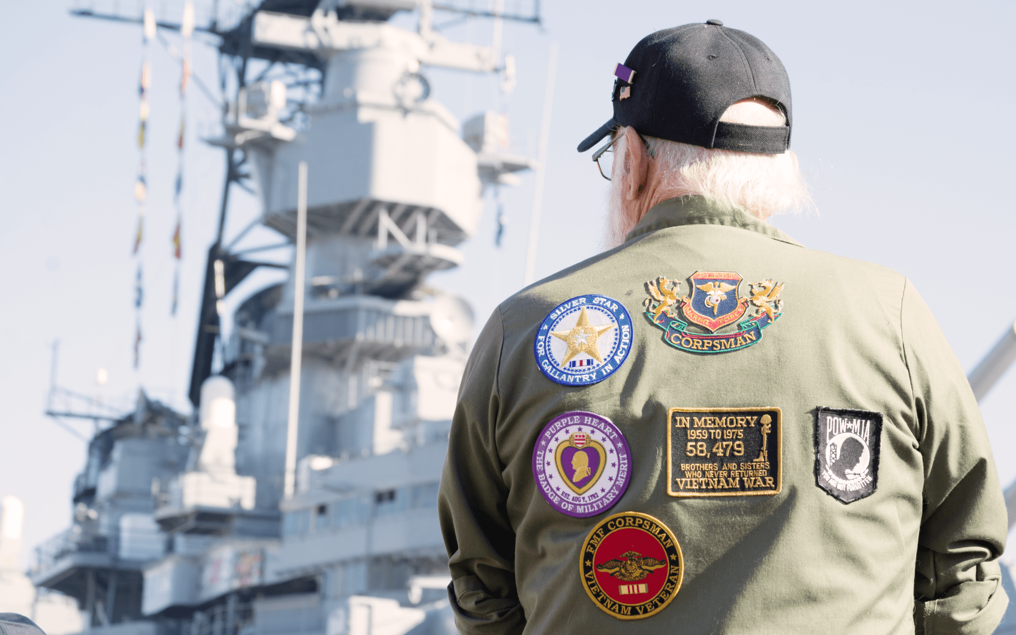 The back of a veteran and his jacket as he stares out at a naval military vessel.