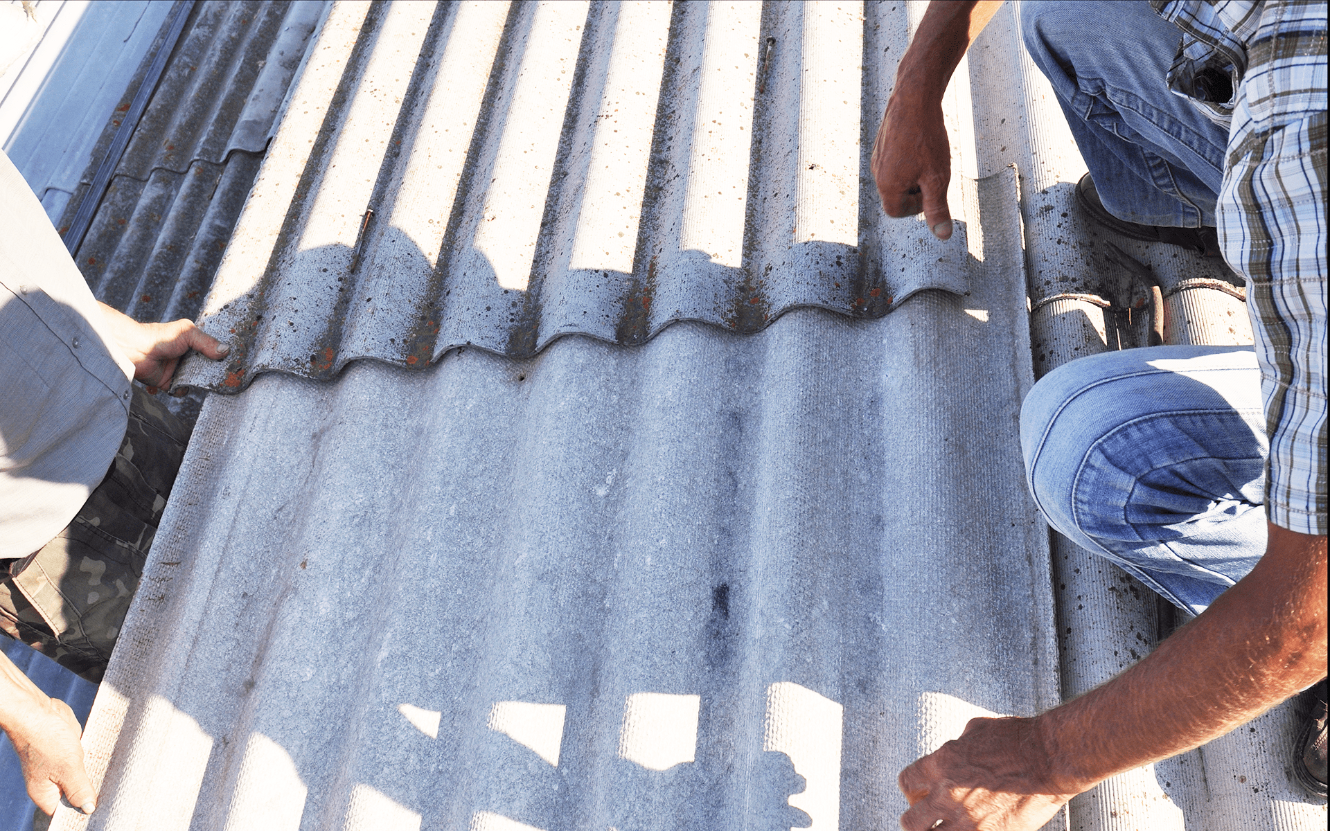 Two men work on asbestos shingles on a roof.