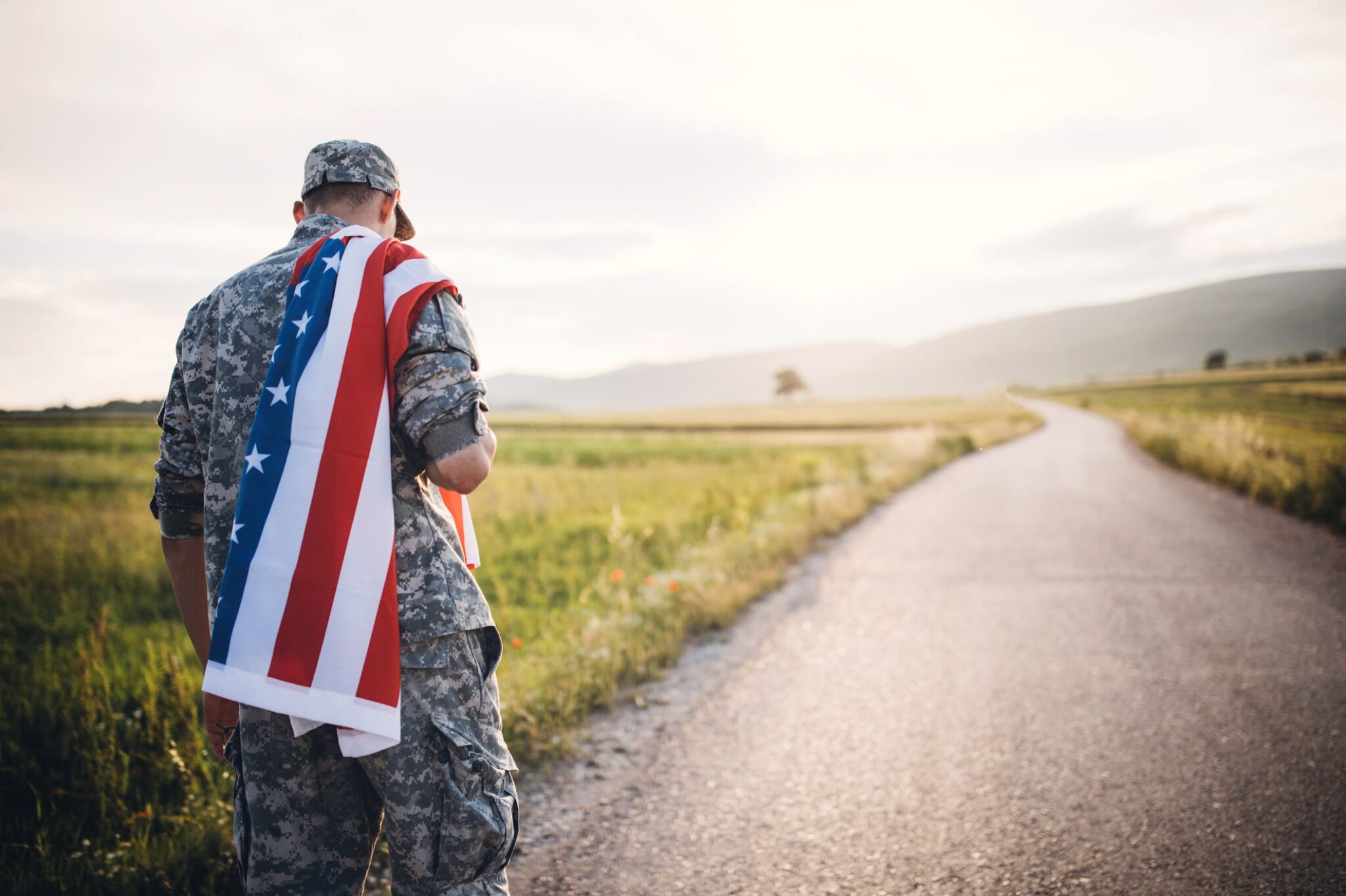 A solider walks down a road alone carrying the U.S Flag on his back.