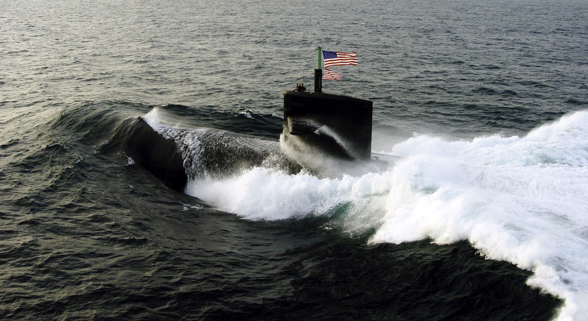 United States Navy submarine comes to the surface of the ocean with the American flag flying on top.