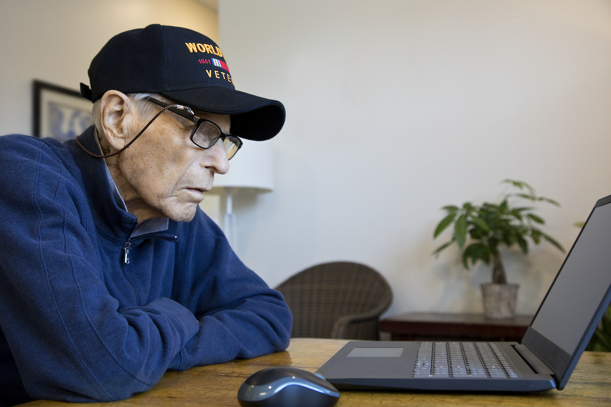 Veteran wearing VFW ball cap focuses on a laptop on the table in fronton him