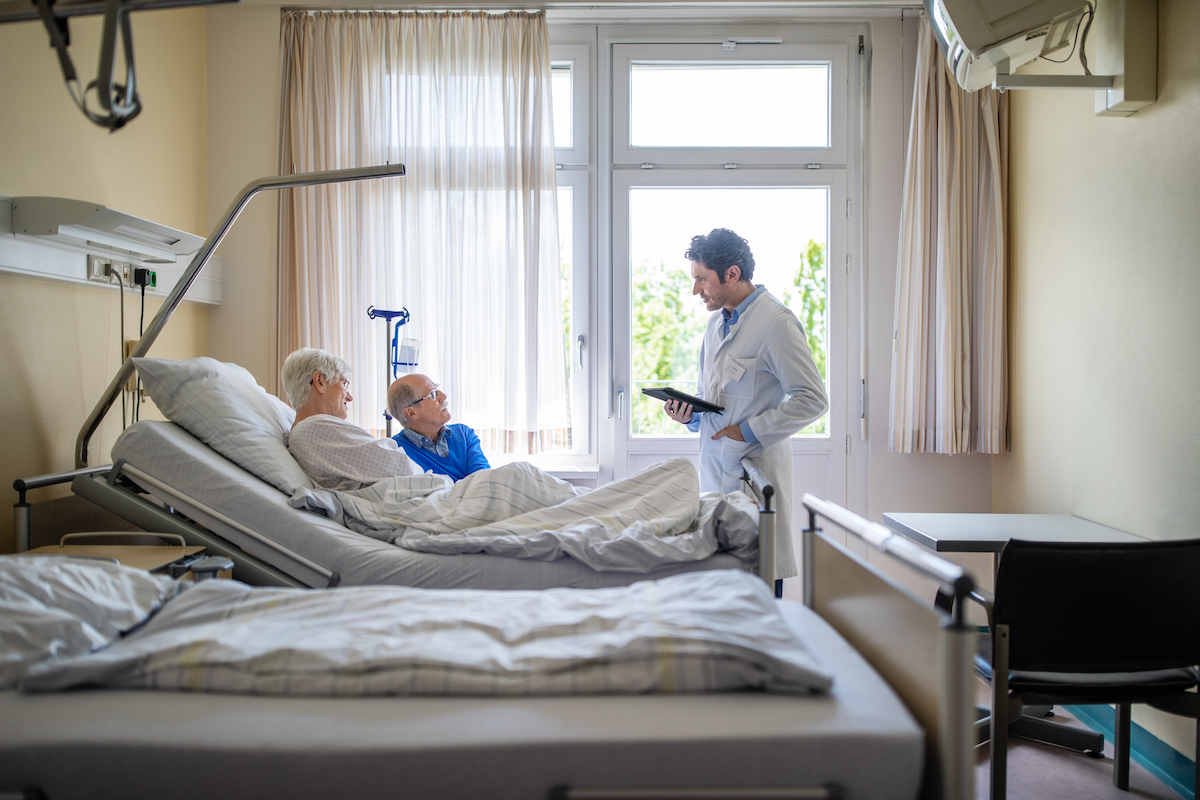 Doctor during a checkup of a senior patient in bed with her partner beside her while at va cancer treatment centers