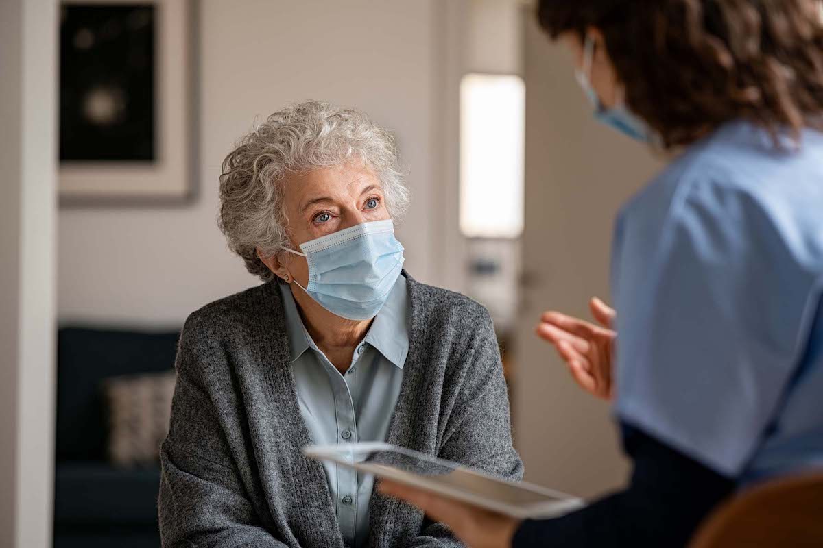 elderly woman talks to her doctor as the doctors holds a tablet device. both wear face masks.