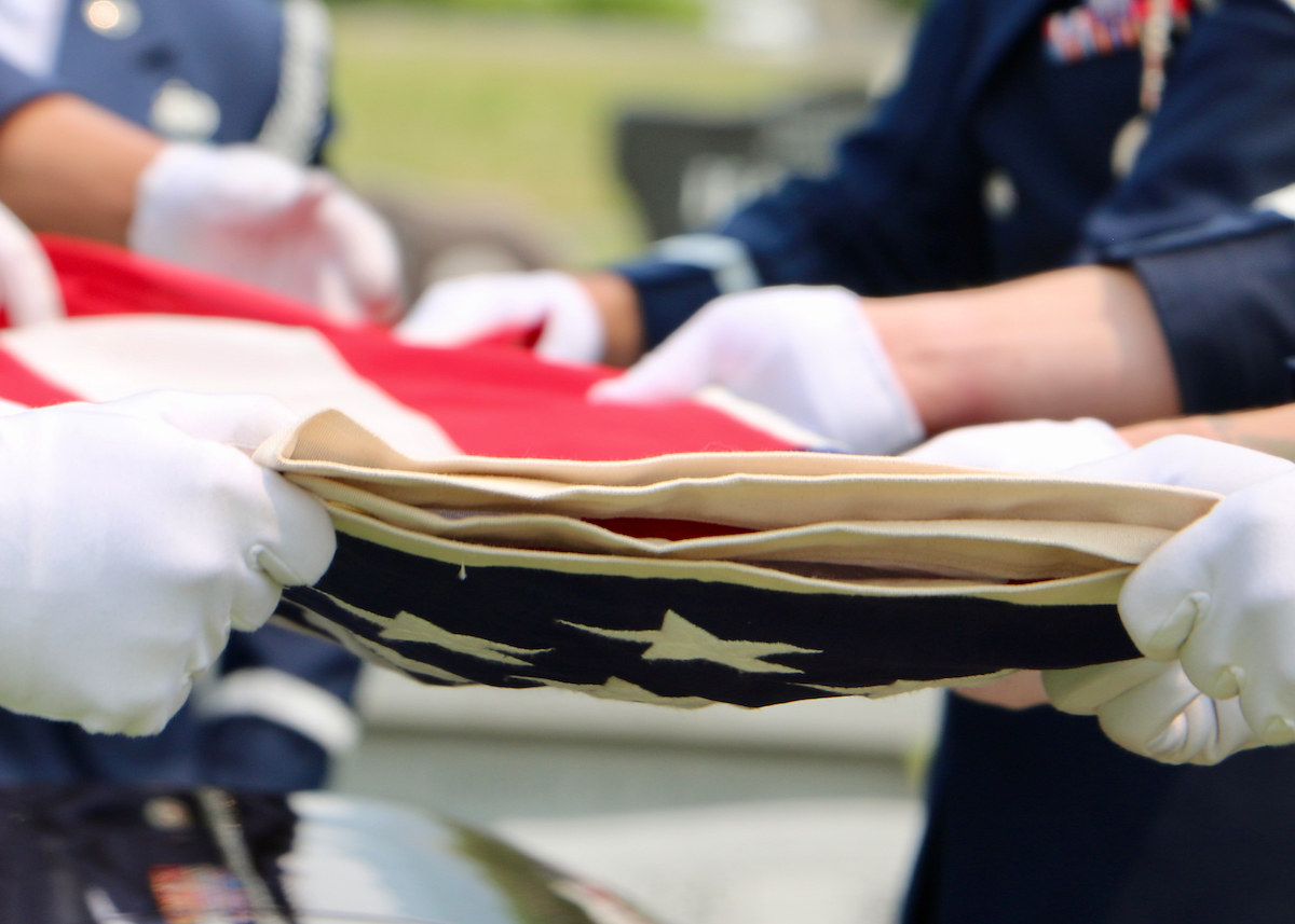 White gloved hands of US Military Honor Guardsmen folding American flag over casket.