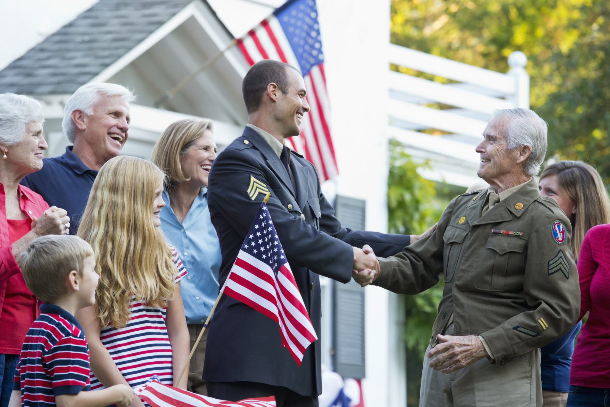 Military man shakes hand with male Veteran in front of house and family with American flags celebrating the passage of the PACT Act