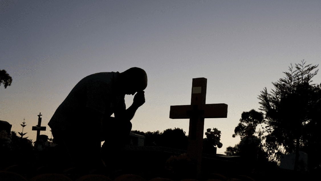 Silhouette of a grieving man at a cemetery beside a grave at sunset. The Grieving Families Act will help families receive compensation on their loved ones behalf.