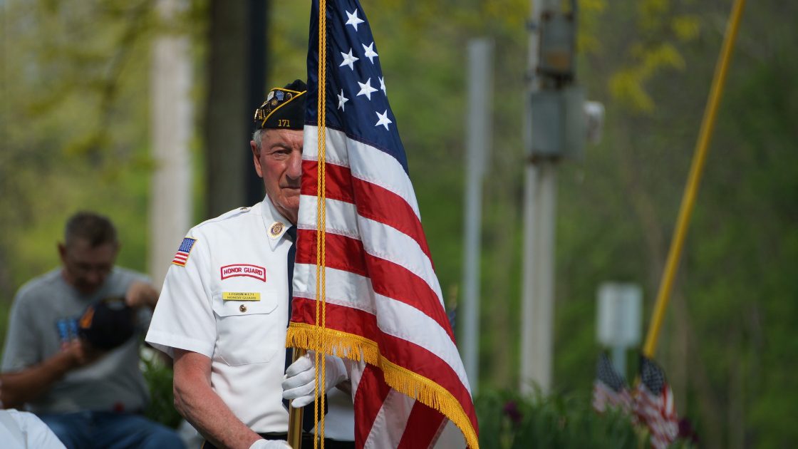 Male Veteran in military outfit holding American flag outside in parade